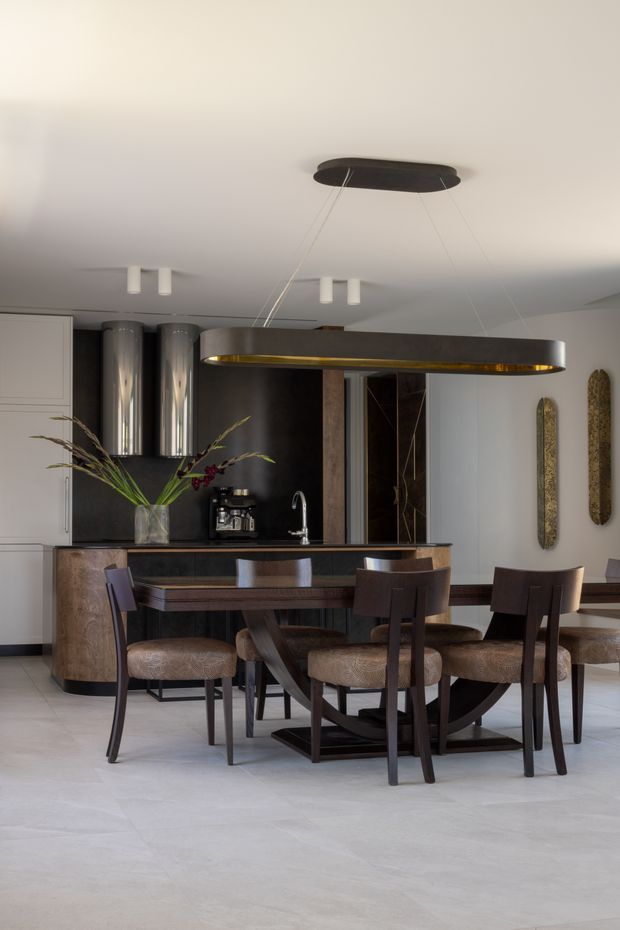 A refined open-plan dining and kitchen interior. A long dark timber dining table with upholstered dining chairs sits beneath a large suspended oval pendant with a dark exterior and warm metallic underside. Behind it is a compact kitchen with dark cabinetry, a stone or dark splashback, integrated appliances, a curved island edge, and a brushed metal tap. The room has pale neutral walls and large light stone-look flooring, with sculptural wall accents and a vase of foliage adding understated decoration. The overall atmosphere is warm, elegant, and contemporary.