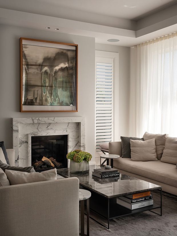 A refined living room with a neutral grey-beige palette, featuring a marble fireplace surround, framed artwork, a large sectional sofa, upholstered armchairs, and a dark glass-topped coffee table. Sheer floor-to-ceiling curtains filter daylight, while plantation shutters appear beside the fireplace. The room has recessed lighting, a tray-like ceiling transition, and polished contemporary furnishings.
