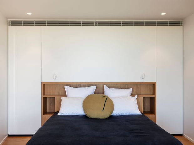 A clean, minimalist bedroom centred on a neatly made bed with white pillows, a dark navy or black coverlet, and a round tan accent cushion. The bed is integrated into warm timber open shelving and cabinetry, with full-height white built-in wardrobes surrounding it. A narrow linear ventilation grille runs above the cabinetry, and recessed ceiling lights provide even illumination. The room has a restrained, contemporary built-in design with warm timber flooring.