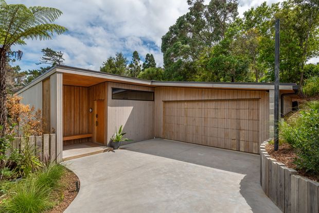 A contemporary single-storey residential garage and entry facade set into a landscaped site. The building has a low-pitched roof, warm vertical timber cladding, a large timber-panel garage door, a recessed entry porch, concrete driveway, dark slender post, and raised garden beds surrounded by mature greenery.