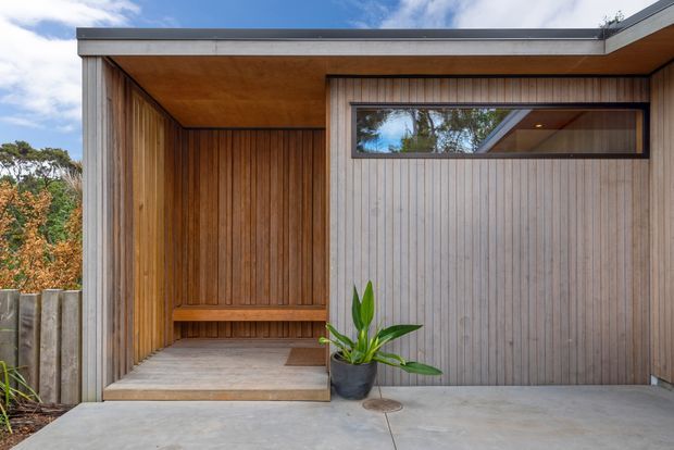 Contemporary timber-clad residential entry facade with a recessed covered porch, vertical timber wall lining, integrated bench seating, a narrow horizontal window, and a flush timber garage or entry door. The structure has a crisp flat roof edge, concrete paving, and a restrained natural palette.