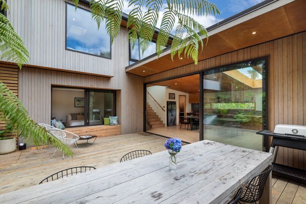 A contemporary timber-clad courtyard and outdoor dining area viewed from a covered patio. The composition features a large weathered timber dining table, black wire dining chairs, a barbecue, timber decking, lush fern planting, and extensive glazing opening into a warm timber-lined interior with a visible stairway and dining area. The home has a sheltered courtyard arrangement, vertical timber wall cladding, and a timber soffit with recessed lighting.