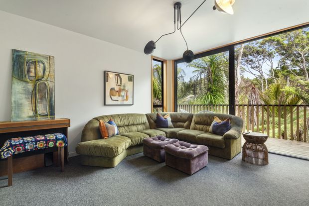 A bright, relaxed living room with a large olive-green sectional sofa, tufted purple ottomans, patterned cushions, a timber console or piano-like piece, framed artwork, and a sculptural suspended light fixture. Full-height corner glazing opens to a timber-railed balcony or deck with lush vegetation beyond. The room has a white ceiling and walls, grey carpet, and warm timber window frames, creating an eclectic, retro-influenced interior with strong indoor-outdoor connection.
