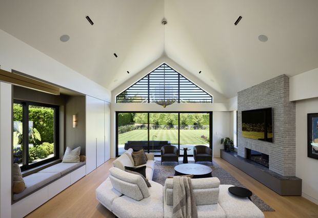Bright contemporary living room with a dramatic vaulted ceiling and triangular clerestory glazing above expansive floor-to-ceiling windows opening to a landscaped garden. The room features a large curved light-coloured sectional sofa, dark lounge chairs, round coffee tables, built-in window seating, timber flooring, recessed lighting, and a textured grey brick fireplace wall with wall-mounted television and low built-in joinery.