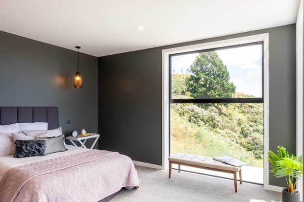 A contemporary bedroom with charcoal grey walls, a large black-framed picture window overlooking greenery, and a light upholstered bed dressed in soft blush and neutral bedding. A slim upholstered bench sits beneath the window, while a warm pendant light and indoor plant add subtle accents.