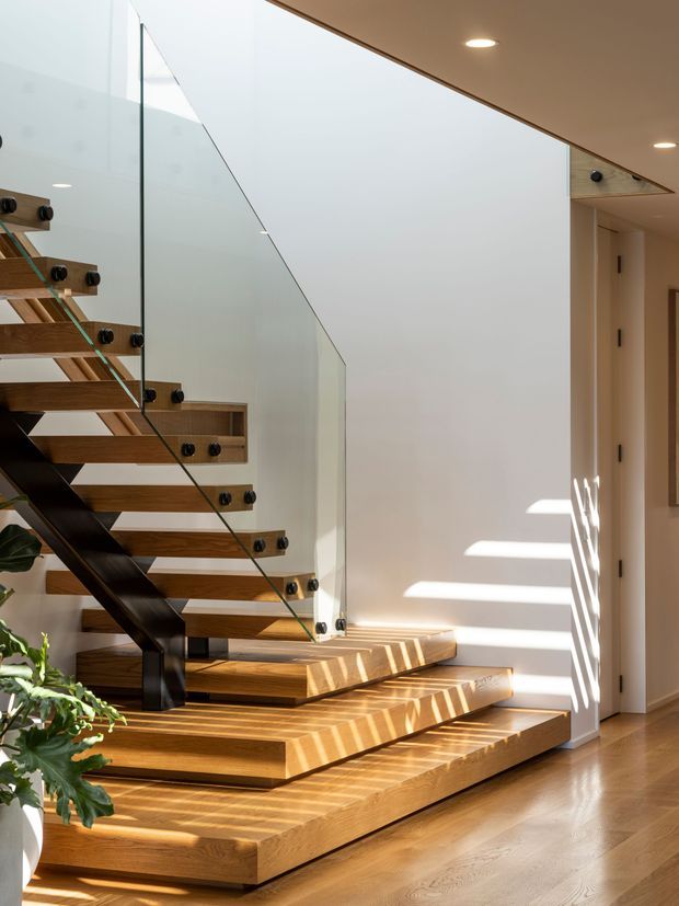 A bright interior stairway with broad floating timber steps and dark structural supports, enclosed by clear glass balustrades with black fittings. The staircase rises beside a white wall, with warm timber flooring, recessed ceiling lights, and strong sunlight casting linear shadows across the steps and wall.