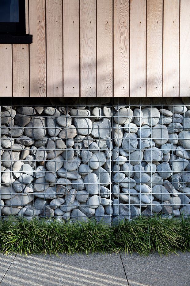 Close-up exterior architectural detail showing pale vertical timber cladding above a gabion-style wall filled with rounded grey river stones in a metal grid. A narrow strip of ornamental grass borders the base, with a concrete or aggregate-paved walkway in the foreground. The image emphasizes natural materials, textural contrast, and a contemporary facade treatment.