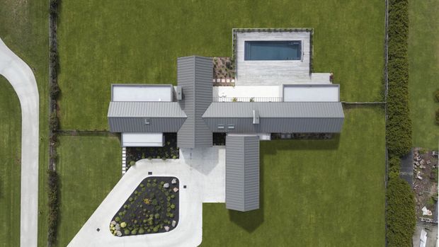 Aerial top-down view of a contemporary cross-shaped residence with multiple grey metal pitched roof volumes, a flat-roofed central section, large paved driveway and entry court, landscaped island, expansive lawn, perimeter hedging, and a rectangular rooftop or courtyard pool.