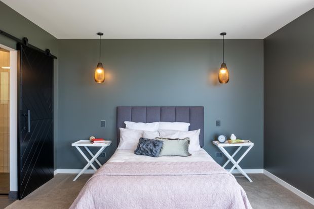 A contemporary bedroom with a centered bed against a dark grey feature wall, upholstered vertical-panel headboard, layered white and pale pink bedding, and symmetrical white X-leg bedside tables. Warm amber pendant lights hang on either side of the bed. A black diagonal-planked barn-style sliding door is visible at left, opening toward an adjoining space. The room has neutral carpet, white skirting, and a clean, balanced composition.