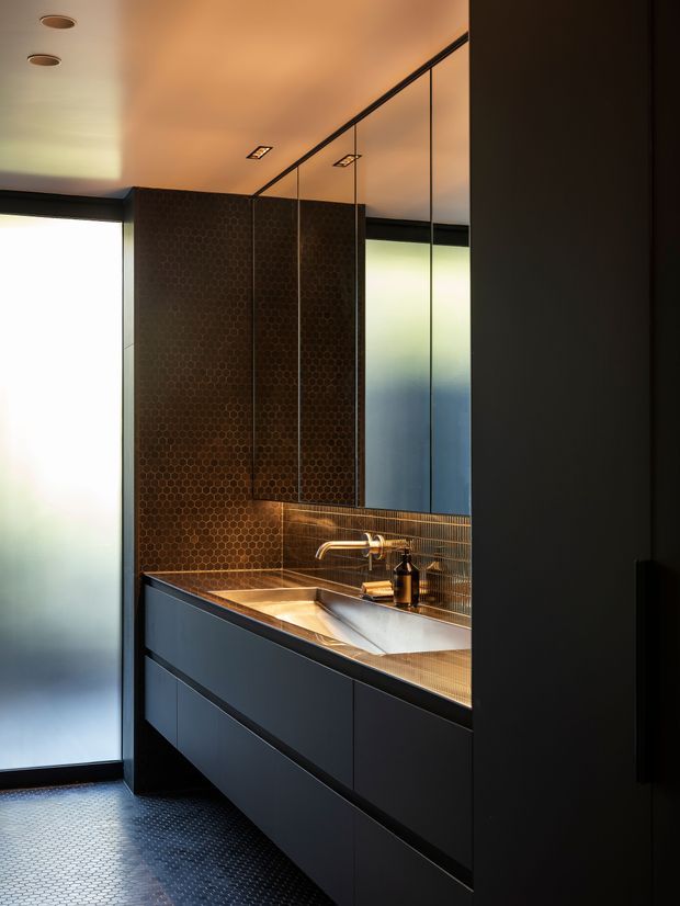 A compact, dark and atmospheric bathroom vanity with a long integrated basin, matte dark cabinetry, bronze-toned honeycomb wall tiles, matching small-format floor tiles, and a reflective mirrored cabinet wall. Frosted glazing at the side brings in diffused light, while warm recessed lighting and metallic tapware create a refined, moody look.