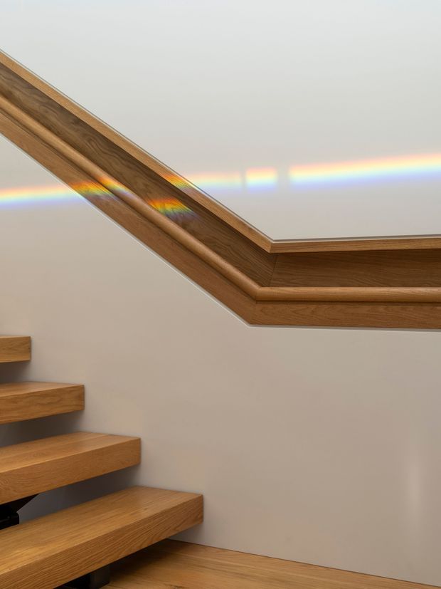 Interior view of a minimalist stairway with floating solid timber treads and matching timber trim around a sloped ceiling or rooflight opening. The warm wood contrasts with smooth light-grey walls, while a rainbow-like light reflection crosses the upper surface.