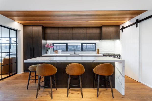 A refined contemporary kitchen with a large dark timber island, white marble-look waterfall benchtop, integrated cabinetry, and three caramel leather bar stools. The room features a dramatic timber slatted ceiling, warm timber flooring, a white tiled splashback, black-framed glazing, and a black barn-style sliding door.