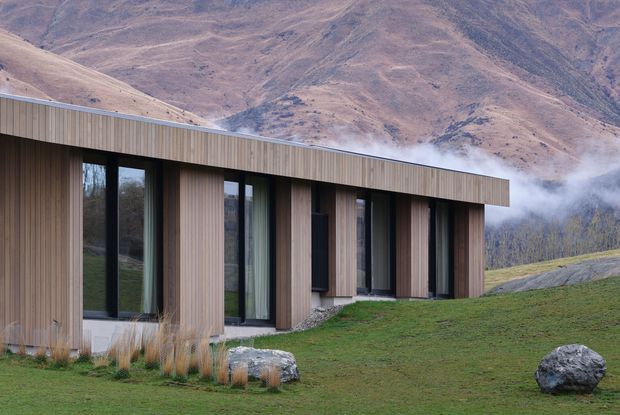 A low, elongated contemporary building set against steep mountainous terrain. The facade combines vertical timber cladding with deep recessed floor-to-ceiling dark-framed glazing, creating a rhythmic series of solid wall bays and windows beneath a simple projecting flat roof. The building sits in a grassy landscaped setting with large natural boulders, while mist and rugged hills form the background.