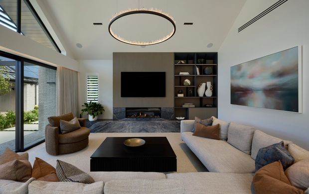 A spacious contemporary living room with a large cream sectional sofa arranged around a black rectangular coffee table, two rounded armchairs, and a dark built-in media wall with an inset fireplace, television, and open shelving. Floor-to-ceiling glazing and a high ceiling bring in abundant daylight, while a large circular pendant light and abstract artwork add visual focus. A landscaped exterior is visible through the glazing.
