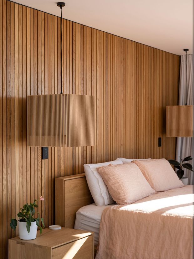 Warm, contemporary bedroom with a full-height vertical timber slat feature wall, matching timber bedside joinery, and two ribbed timber pendant lamps. The bed is dressed in soft blush and white linens, with a potted plant and curtains adding a relaxed, natural feel. Strong sunlight creates a bright, calm atmosphere.