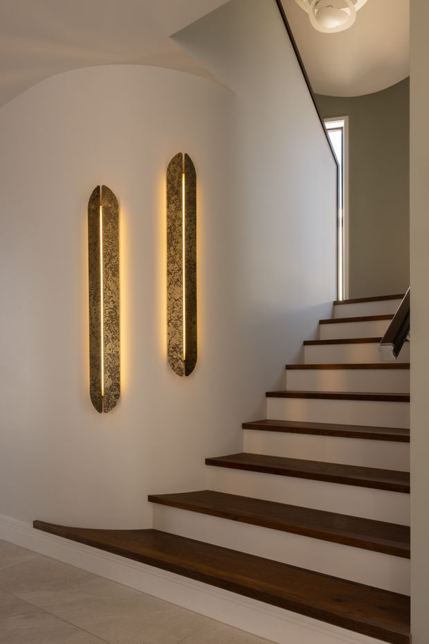 A refined interior stairway with dark timber stair treads and a white painted stair stringer and walls. Two tall, narrow decorative wall sconces with patterned metallic faces provide warm illumination, while a sculptural circular ceiling light and a slim vertical window add architectural interest. Pale tiled flooring is visible at the base of the stairs.