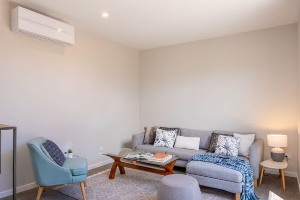 Bright, compact living room with a grey upholstered sofa, patterned cushions, blue throw, glass-topped timber coffee table, round ottoman, pale blue armchair, side tables and soft neutral walls. A wall-mounted split-system air conditioner and recessed ceiling light are visible.