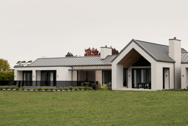 Single-storey contemporary residence viewed from the front across a broad lawn. The home has white rendered walls, dark standing-seam metal roofing, strong gabled forms, projecting chimney elements, extensive dark-framed glazing, and a prominent covered entry or outdoor living void with a timber-lined soffit.