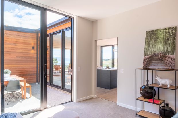 Bright contemporary interior with a neutral carpeted floor and white walls, opening through large black-framed glazed doors to a timber-clad courtyard or terrace. A compact dark kitchen is visible through the internal opening, while a slim black metal display shelf and woodland artwork furnish the foreground.