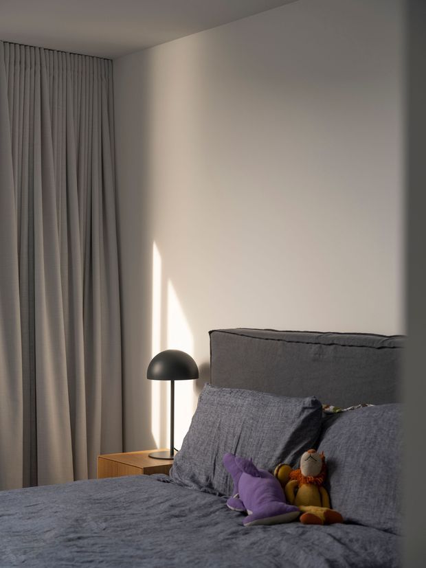 A softly lit, minimalist bedroom with a large upholstered grey bedhead, grey textured bedding and pillows, full-height pleated curtains, and a small timber bedside table with a black dome lamp. Plush toys rest on the bed, adding a subtle playful touch. Warm sunlight creates a narrow highlight across the neutral wall.