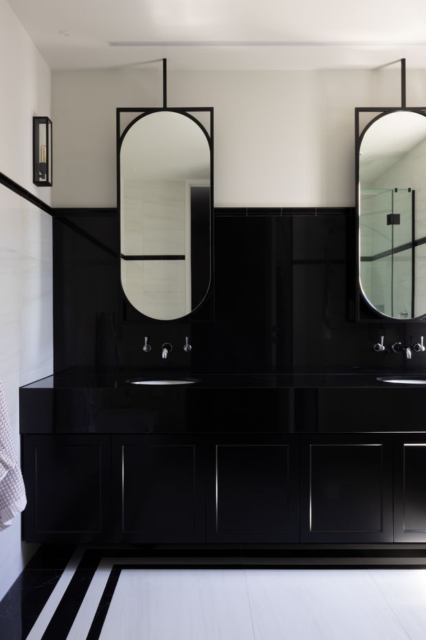 A sophisticated black-and-white bathroom featuring a long black vanity with integrated basins, black cabinetry, dark wall tiling, chrome wall-mounted tapware, and two tall arched mirrors in black frames suspended from the ceiling. White upper walls and a geometric black-and-white floor border create a bold, high-contrast scheme.