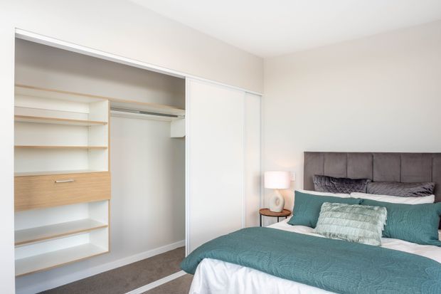 Bright, compact bedroom with a large built-in wardrobe and open shelving along the left wall. The room features a upholstered charcoal-grey bedhead, teal bedding and cushions, a small timber bedside table, and a warm bedside lamp. White walls and cabinetry create a clean, contemporary appearance, with grey carpet underfoot.