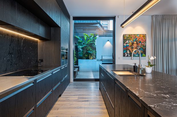 Contemporary galley-style kitchen with dark timber cabinetry, a large marble-look island, integrated linear lighting, and full-height glazing opening to a lush courtyard or outdoor area. A colourful artwork, pendant lighting, and sheer curtains add warmth and contrast to the refined kitchen interior.