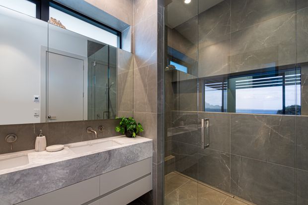 Contemporary bathroom with a double vanity featuring a pale stone benchtop and streamlined white drawers. Large grey stone-look wall and floor tiles continue into a frameless glass shower enclosure. A wide horizontal window provides natural light and views beyond, while dark window and shower hardware add contrast.