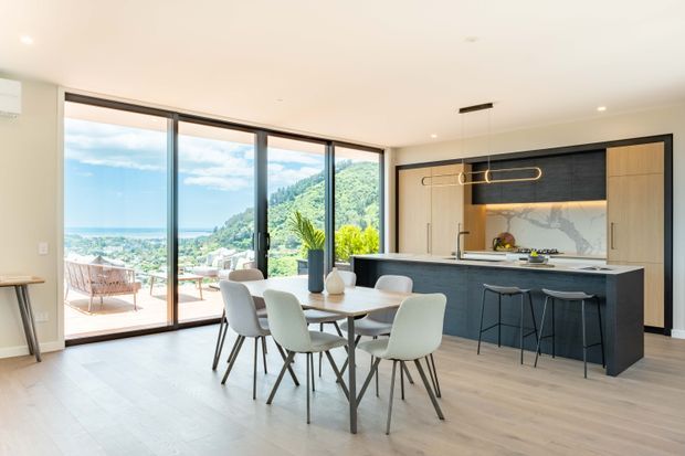 Bright open-plan kitchen and dining area with floor-to-ceiling sliding glazing opening onto a terrace and expansive coastal hillside view. The space features pale timber flooring, a dark kitchen island with integrated seating, light wood cabinetry, a marble-look splashback, contemporary dining furniture, and sculptural linear pendant lighting.