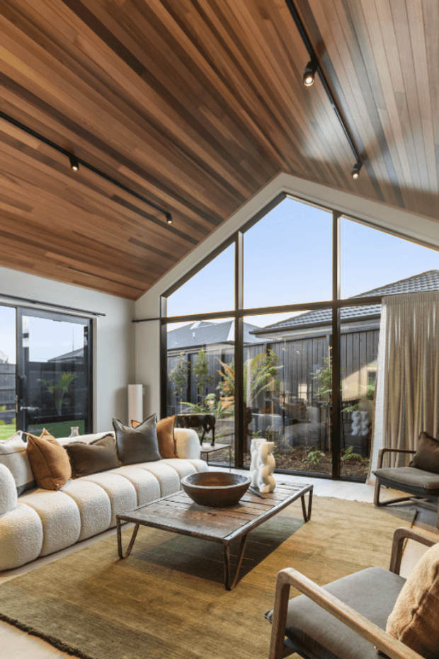 A warm, contemporary living room with a large cream boucle sectional, timber-framed armchairs, a low rustic coffee table, and a muted tan area rug. The room features extensive floor-to-ceiling glazing overlooking a planted courtyard, along with a dramatic sloped ceiling clad in warm timber boards and black track lighting. Neutral furnishings and natural materials create a relaxed, refined atmosphere.