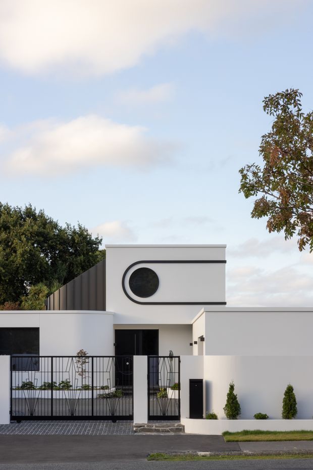Street-facing view of a contemporary white residential facade with strong geometric forms, a distinctive circular porthole-style window outlined in dark trim, vertical dark metal side cladding, and a black metal front fence and gate. The home has a low, flat roofline and minimalist landscaping along the frontage.