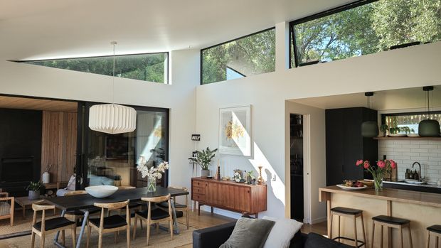 Bright open-plan contemporary living, dining and kitchen interior with a high ceiling and extensive clerestory glazing. The room features a dark timber and black-framed glazed partition, a timber dining table with chairs, a mid-century-inspired sideboard, pale walls, dark kitchen cabinetry, a light stone or solid-surface island, and a white rectangular-tile splashback. Sunlight and views of mature trees are visible through the windows.