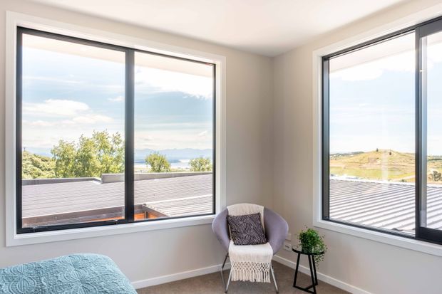 Bright bedroom corner with large black-framed windows overlooking a coastal or hilly landscape. The room has light neutral walls, a partially visible bed with a textured blue cover, and a modern accent chair with a woven throw beside a small black side table and plant.