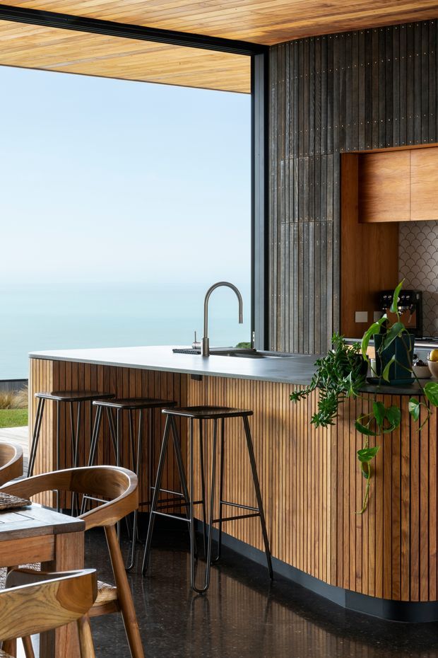 A refined kitchen and dining area with a long island facing expansive ocean views. The kitchen features vertical timber-panelled cabinetry, a dark stone benchtop, black-framed floor-to-ceiling glazing, a timber-lined ceiling, dark vertical wall cladding, and a decorative scallop-pattern splashback. Black bar stools line the island, while timber dining furniture and indoor plants occupy the foreground.