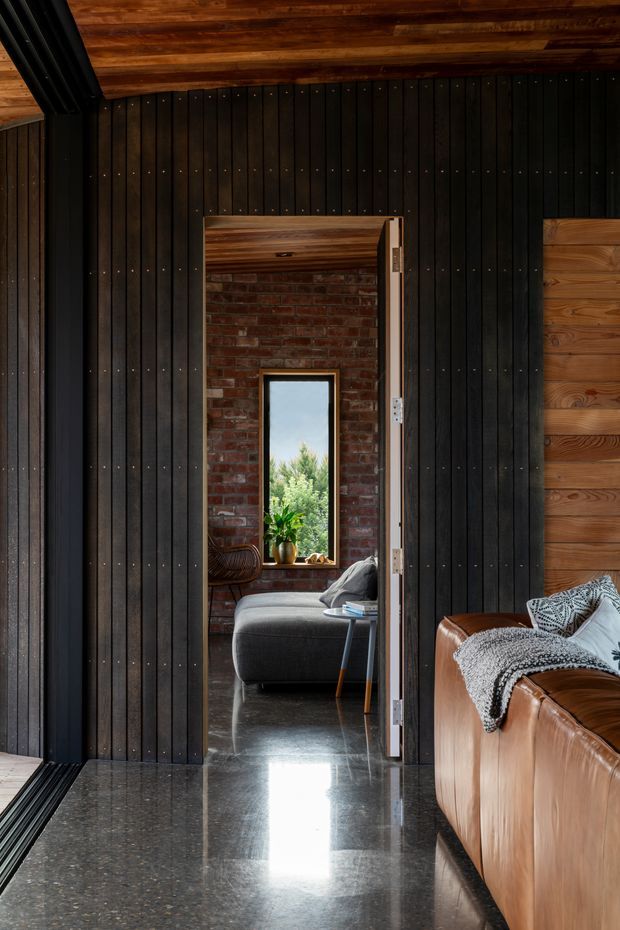 Interior view of a dark, refined modern living space framed by black vertical timber wall panelling and a warm timber ceiling. A polished dark stone or concrete floor leads through an open doorway into a compact lounge with a grey sofa, exposed red-brick wall, and narrow garden-facing window. A tan leather sofa with textured throws occupies the foreground.