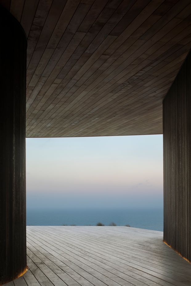 A dramatic architectural view through a tall, open-sided passage or pavilion toward the ocean. Dark vertically textured wall elements frame a broad timber-planked deck, while matching timber boards form the overhead soffit and create a continuous, sculptural enclosure. The minimal composition emphasizes the expansive coastal horizon and strong indoor-outdoor connection.