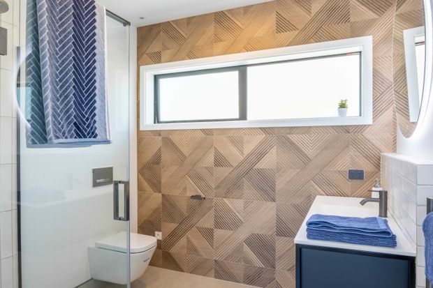 Bright contemporary bathroom featuring a frameless glass shower enclosure, wall-hung toilet, floating dark vanity, and a long horizontal window. The main feature is a geometric chevron-pattern timber wall finish in warm light wood tones, complemented by blue towels and matte dark hardware.