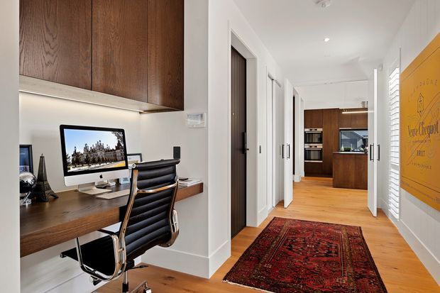 A bright, modern residential hallway with a compact built-in home office on the left. The office features a floating dark timber desk, integrated dark timber overhead cabinetry, computer workstation, and black leather office chair. The hallway has white walls and trim, warm timber flooring, multiple internal doors, a red patterned runner rug, and a kitchen visible at the far end. A large mustard-yellow framed artwork adds a bold accent on the right wall.