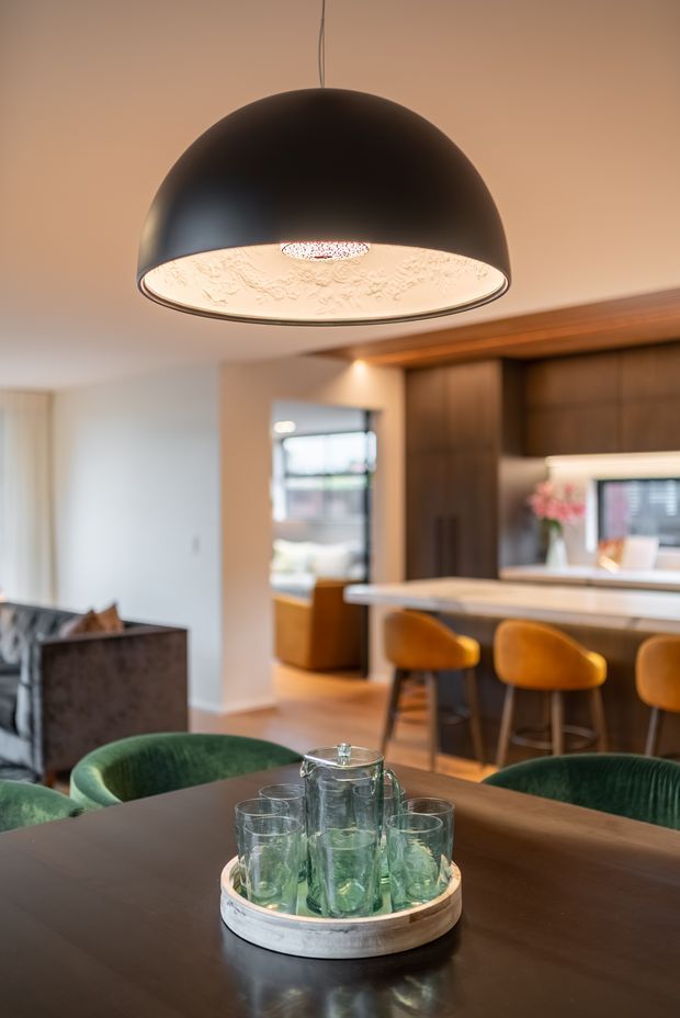 Warm contemporary open-plan dining and kitchen interior, viewed from a dark timber dining table. A large black dome pendant light dominates the foreground, with green upholstered dining chairs, a round tray and glassware on the table, and a kitchen island with timber bar stools and dark wood cabinetry in the background.