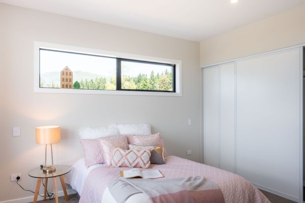 Bright, compact bedroom with a soft neutral palette, a bed dressed in blush pink and white bedding, patterned cushions, a small glass-topped bedside table with a lamp, built-in white sliding-door wardrobe, and a wide horizontal window overlooking greenery and distant hills.