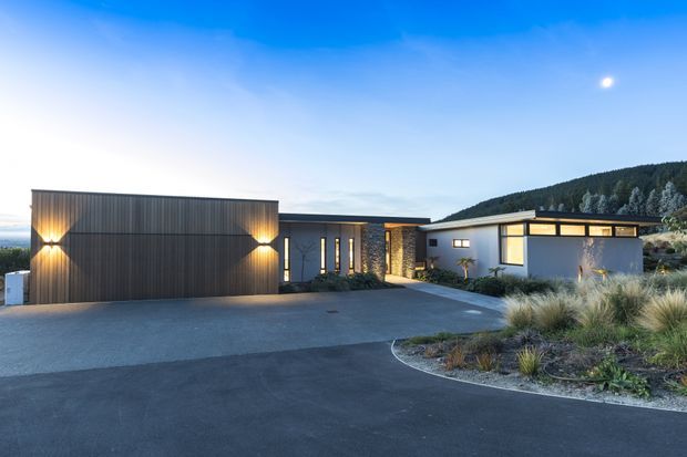 A contemporary single-storey residence photographed at dusk, with a broad dark vertical-metal-clad garage volume, warm exterior wall lighting, a recessed glazed entry, and a low flat-roofed white wing. The home is set on a gently landscaped site with ornamental grasses and a paved driveway, with a wooded hillside visible behind it.