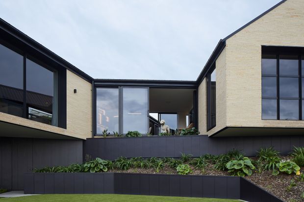 Exterior view of a contemporary courtyard-style residence with pale cream brick walls, large black-framed glazing, dark vertical cladding and layered raised garden beds. The central recessed opening creates a sheltered courtyard or terrace, with landscaped planting in the foreground and a sculptural figure visible through the glazing.