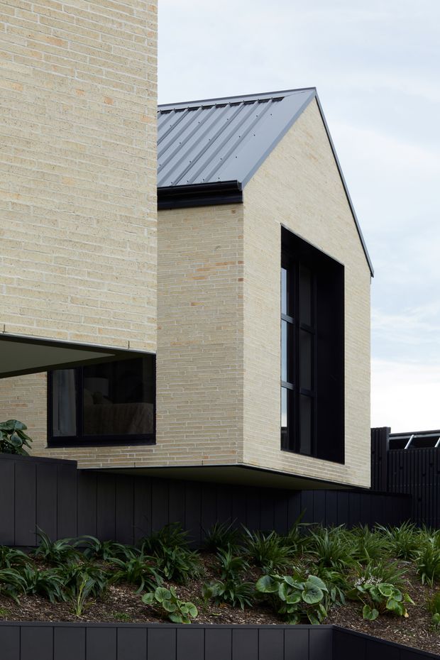 Exterior architectural photograph of a contemporary two-storey home with pale yellow brick walls, a dark grey standing-seam gabled metal roof, large black-framed windows, and a projecting upper volume. Dark vertical fencing and landscaped garden beds occupy the foreground.