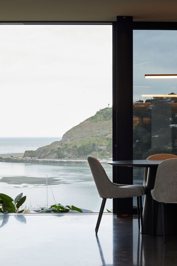 A refined dining area beside expansive floor-to-ceiling glazing, overlooking a calm coastal inlet and green headland. The space features a small dark round dining table, upholstered dining chairs, glossy dark flooring, black-framed glazing, and slim linear pendant lighting. Indoor plants sit near the window, with strong natural light and a serene waterfront outlook.