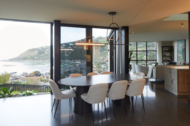 Contemporary open-plan dining and living area with a large round dining table and upholstered chairs beside expansive floor-to-ceiling glazing. The windows overlook a bright coastal harbour or inlet setting, while a modern kitchen and lounge area sit beyond the dining space. Dark polished flooring, black window framing, warm timber cabinetry, and sculptural pendant lighting create a refined, high-contrast interior.