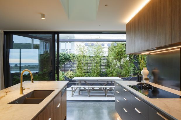 Contemporary open-plan kitchen with a large central island, integrated double sink, timber cabinetry, dark lower drawers, pale stone benchtops and warm under-cabinet lighting. Full-height glazing opens toward an outdoor dining courtyard with a picnic-style table, while a coastal view is visible through the side glazing.