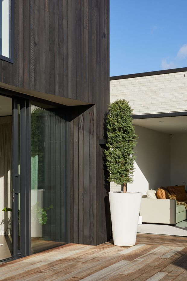 A contemporary residential courtyard or terrace viewed from outside, featuring dark vertical timber cladding, large glazed doors, pale horizontal brickwork, and a timber deck. An open living area is visible through the glazing, with a tall potted tree and outdoor seating creating a refined indoor-outdoor connection.