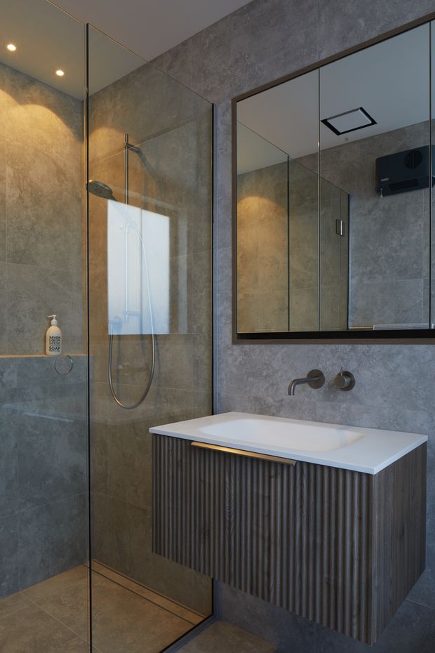 Contemporary bathroom featuring a frameless glass walk-in shower, large grey stone-look wall and floor tiles, a wall-mounted fluted dark timber vanity with white integrated basin, brushed metal wall-mounted tapware, and a large black-framed mirrored cabinet. Warm recessed lighting creates a refined, moody atmosphere.