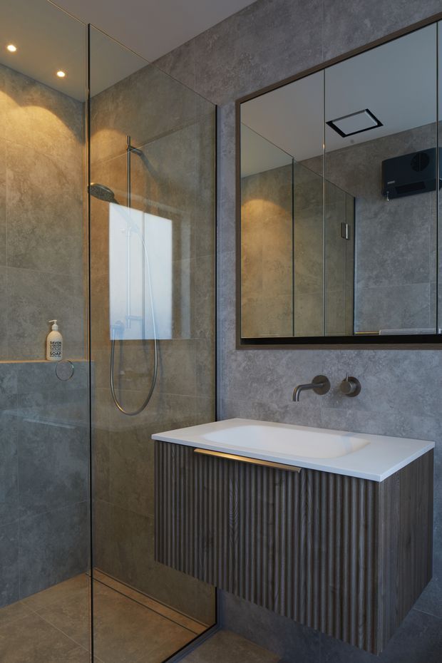A contemporary bathroom featuring a frameless glass walk-in shower, grey stone-look wall and floor finishes, a wall-mounted fluted dark vanity with a white integrated basin, brushed metal tapware, and a large black-framed mirrored cabinet. Recessed ceiling lighting and warm under-vanity illumination create a refined, moody atmosphere.