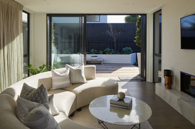 Bright contemporary living room with a curved cream sectional sofa, textured cushions, round marble coffee table, wall-mounted television and low built-in media cabinetry. Full-height glazed sliding doors open directly to a private landscaped courtyard with raised planters and outdoor steps.